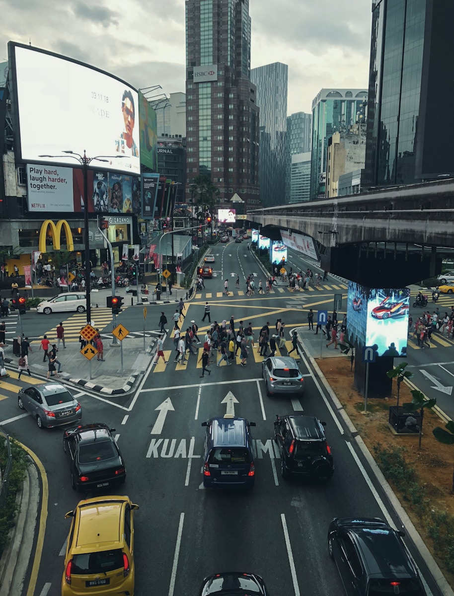 people walking on pathway near buildings and different vehicles on road under white and gray sky during daytime