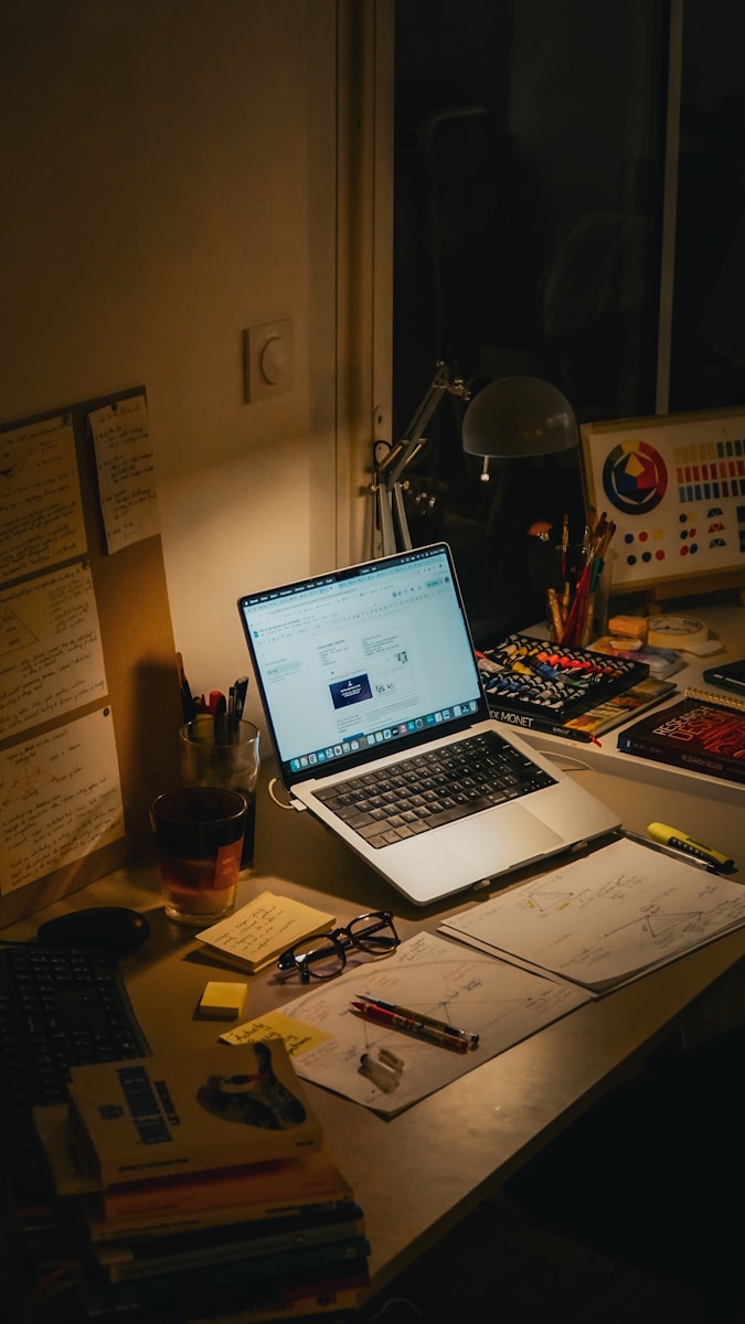 Desk with laptop, books, and papers at night.