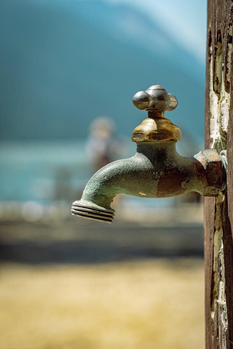 Close-up of a rusty faucet outdoors, set against a blurred lake and mountain background, evoking a nostalgic summer feel.