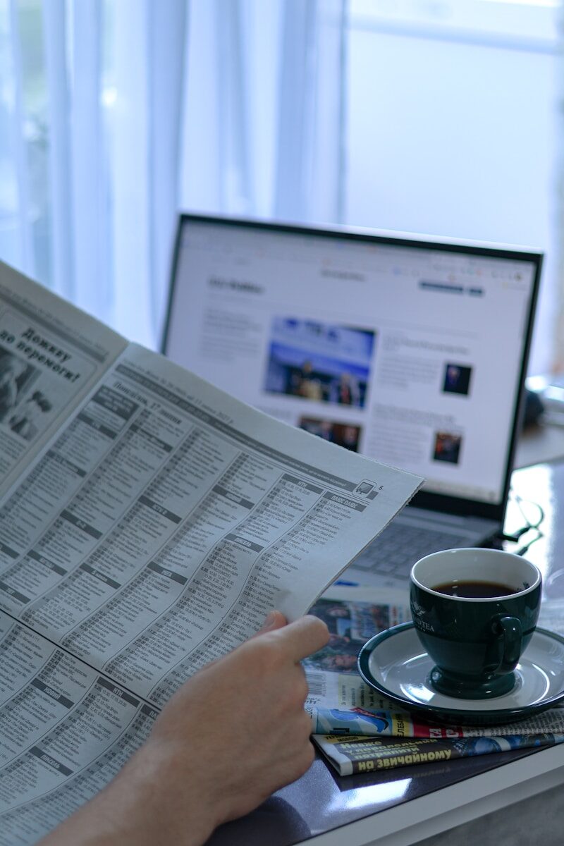 a person reading a newspaper next to a laptop
