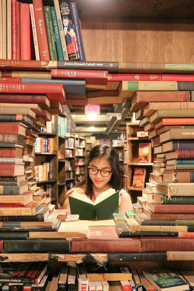 A woman with glasses reading a book framed by stacks of old books